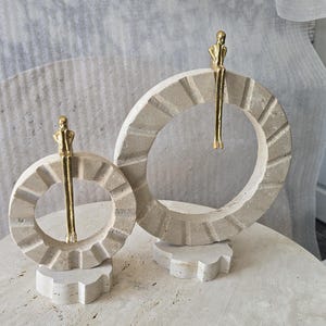 Pu&ograve; includere: Due sculture circolari in pietra con una finitura beige testurizzata, ognuna caratterizzata da una piccola figura metallica color oro appollaiata sul bordo superiore con le gambe a penzoloni. Le sculture sono montate su basi in pietra.
