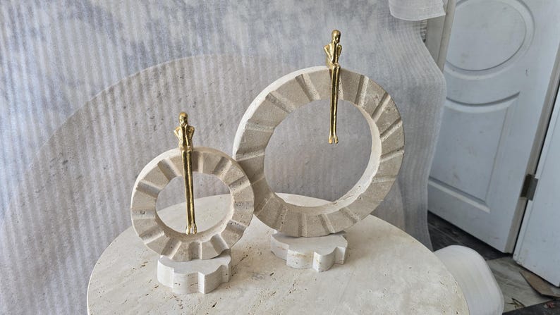 Pu&ograve; includere: Due sculture circolari in pietra con una finitura beige testurizzata, ognuna caratterizzata da una piccola figura metallica color oro appollaiata sul bordo superiore con le gambe a penzoloni. Le sculture sono montate su basi in pietra.