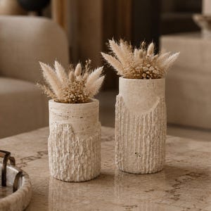 May include: Two cylindrical off-white stone vases with a textured, rugged lower section and a smooth upper rim. Each vase contains a decorative arrangement of dried beige grasses and small fluffy plumes, creating a neutral, earthy aesthetic.