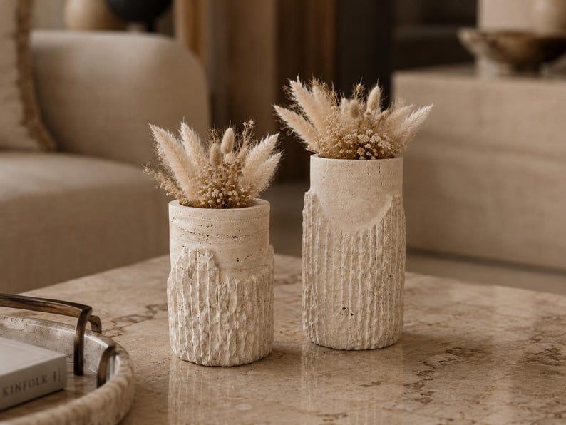 May include: Two cylindrical off-white stone vases with a textured, rugged lower section and a smooth upper rim. Each vase contains a decorative arrangement of dried beige grasses and small fluffy plumes, creating a neutral, earthy aesthetic.