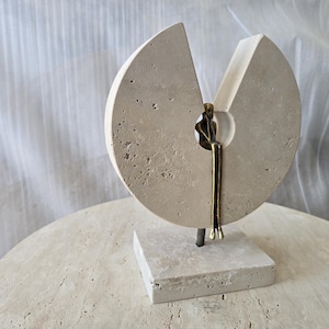 K&ouml;nnte beinhalten: Eine Skulptur bestehend aus einer kreisf&ouml;rmigen, por&ouml;sen beigen Steinscheibe mit keilf&ouml;rmigem Ausschnitt, montiert auf einem quadratischen Steinsockel. Eine kleine, bronzefarbene Metallfigur sitzt in der zentralen &Ouml;ffnung mit baumelnden Beinen.