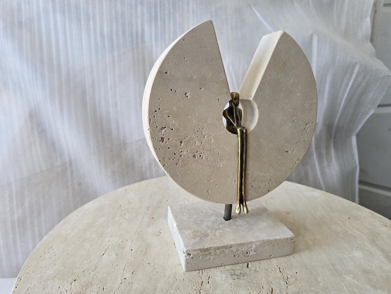 K&ouml;nnte beinhalten: Eine Skulptur bestehend aus einer kreisf&ouml;rmigen, por&ouml;sen beigen Steinscheibe mit keilf&ouml;rmigem Ausschnitt, montiert auf einem quadratischen Steinsockel. Eine kleine, bronzefarbene Metallfigur sitzt in der zentralen &Ouml;ffnung mit baumelnden Beinen.