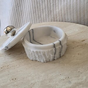 May include: A round white marble container with grey veining and a textured exterior. The matching lid features a smooth, polished silver-toned spherical knob and is shown resting partially off the top of the container, revealing the interior.