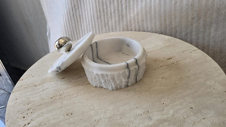 May include: A round white marble container with grey veining and a textured exterior. The matching lid features a smooth, polished silver-toned spherical knob and is shown resting partially off the top of the container, revealing the interior.