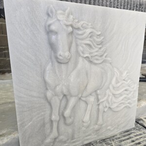 May include: A square white marble slab featuring a high-relief carving of a horse in motion. The horse is depicted facing forward with a flowing mane and tail, set against a textured background that mimics the appearance of wind or movement.