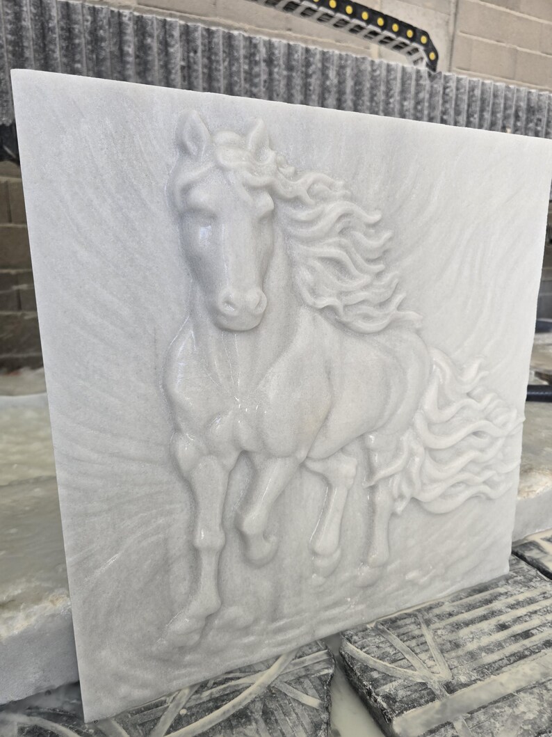 May include: A square white marble slab featuring a high-relief carving of a horse in motion. The horse is depicted facing forward with a flowing mane and tail, set against a textured background that mimics the appearance of wind or movement.