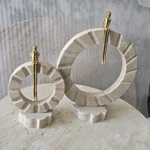 Pu&ograve; includere: Due sculture circolari in pietra con una superficie beige testurizzata e scanalata. Ogni anello presenta una piccola figura metallica color oro appollaiata sul bordo superiore, con le gambe che penzolano al centro dell'apertura circolare.