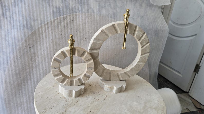 Pu&ograve; includere: Due sculture circolari in pietra con una superficie beige testurizzata e scanalata. Ogni anello presenta una piccola figura metallica color oro appollaiata sul bordo superiore, con le gambe che penzolano al centro dell'apertura circolare.