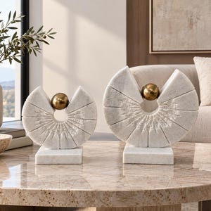 May include: Two white marble sculptures featuring a semi-circular fan design with radial grooves and a central metallic gold sphere. Each piece is mounted on a rectangular white marble base, showcasing a smooth finish and textured detailing.