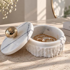 May include: A round white marble jewellery box with a textured exterior and a smooth lid featuring a gold-toned hammered metal sphere handle. The lid is tilted to the side, revealing a gold chain necklace and a ring resting inside the container.