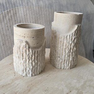 May include: Two cylindrical beige stone vases with a rough, textured exterior resembling tree bark on the lower half and a smooth, layered finish on the upper rim. The vases are crafted from natural travertine stone with visible porous details.