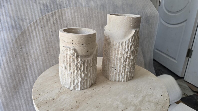 May include: Two cylindrical beige stone vases with a rough, textured exterior resembling tree bark on the lower half and a smooth, layered finish on the upper rim. The vases are crafted from natural travertine stone with visible porous details.