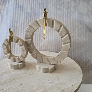 Pu&ograve; includere: Due sculture decorative composte da anelli circolari in pietra beige con bordi dentellati, montati su una piccola base. Una piccola figura umana astratta di colore dorato &egrave; seduta sulla parte superiore di ogni anello con le gambe a penzoloni.