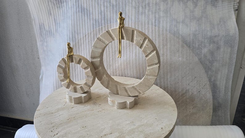 Pu&ograve; includere: Due sculture decorative composte da anelli circolari in pietra beige con bordi dentellati, montati su una piccola base. Una piccola figura umana astratta di colore dorato &egrave; seduta sulla parte superiore di ogni anello con le gambe a penzoloni.