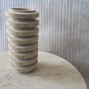 May include: A cylindrical beige travertine vase featuring a series of stacked, horizontal circular discs along its exterior. The stone surface displays a natural porous texture with subtle variations in colour, creating a layered, ribbed appearance.