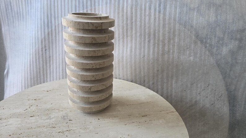 May include: A cylindrical beige travertine vase featuring a series of stacked, horizontal circular discs along its exterior. The stone surface displays a natural porous texture with subtle variations in colour, creating a layered, ribbed appearance.