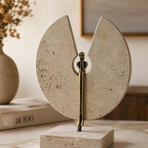 K&ouml;nnte beinhalten: Eine dekorative Skulptur bestehend aus einer kreisf&ouml;rmigen, por&ouml;sen beigen Steinscheibe mit einem keilf&ouml;rmigen Ausschnitt oben. Eine schlanke, bronzefarbene Metallfigur sitzt in der zentralen &Ouml;ffnung, mit langen Beinen, die zum quadratischen Steinsockel f&uuml;hren.