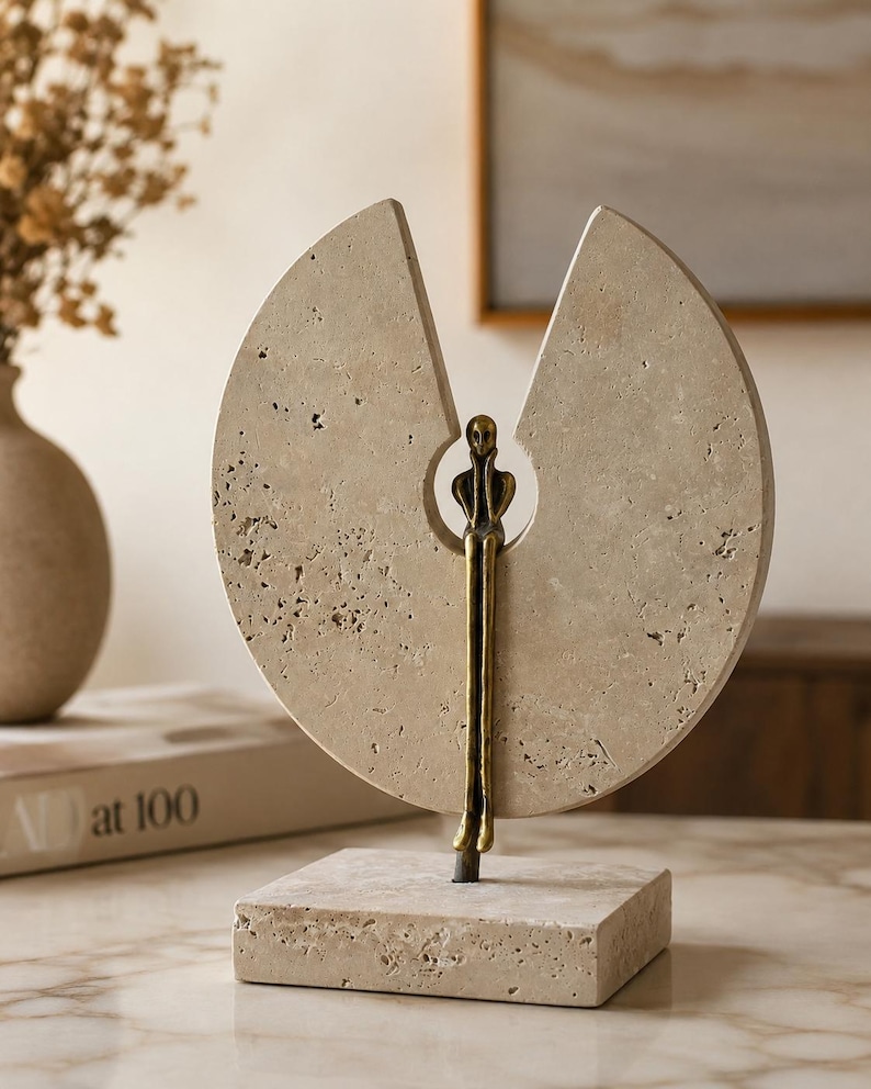 K&ouml;nnte beinhalten: Eine dekorative Skulptur bestehend aus einer kreisf&ouml;rmigen, por&ouml;sen beigen Steinscheibe mit einem keilf&ouml;rmigen Ausschnitt oben. Eine schlanke, bronzefarbene Metallfigur sitzt in der zentralen &Ouml;ffnung, mit langen Beinen, die zum quadratischen Steinsockel f&uuml;hren.