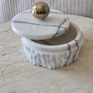 May include: A round white marble container with black veining, featuring a textured, chiseled exterior finish. The lid is topped with a smooth, polished gold-toned spherical handle, creating a contrast between the rough stone and the metallic accent.