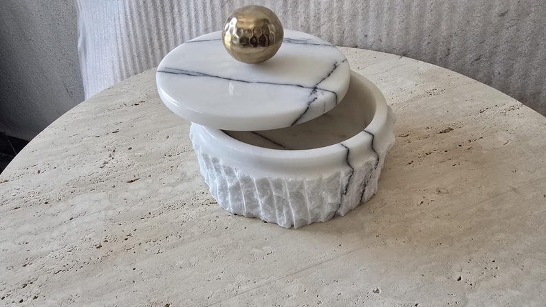 May include: A round white marble container with black veining, featuring a textured, chiseled exterior finish. The lid is topped with a smooth, polished gold-toned spherical handle, creating a contrast between the rough stone and the metallic accent.