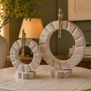 Pu&ograve; includere: Due sculture circolari in pietra con finitura beige testurizzata, ciascuna caratterizzata da una piccola figura metallica color oro seduta sul bordo superiore. Le sculture poggiano su piccole basi in pietra a forma di ingranaggio.