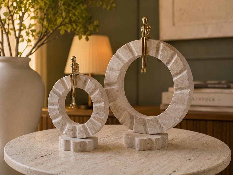 Pu&ograve; includere: Due sculture circolari in pietra con finitura beige testurizzata, ciascuna caratterizzata da una piccola figura metallica color oro seduta sul bordo superiore. Le sculture poggiano su piccole basi in pietra a forma di ingranaggio.