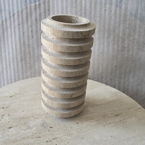 May include: A cylindrical vase crafted from beige travertine stone, featuring a series of stacked, horizontal circular ridges along its exterior. The surface of the stone displays natural porous textures and variations in colour throughout the structure.