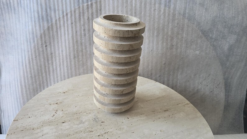 May include: A cylindrical vase crafted from beige travertine stone, featuring a series of stacked, horizontal circular ridges along its exterior. The surface of the stone displays natural porous textures and variations in colour throughout the structure.