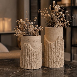 May include: Two cylindrical beige stone vases with a rough, vertically grooved texture on the lower half and a smooth upper section. Each vase contains a bouquet of dried, light-colored flowers with small, delicate, clustered blossoms.