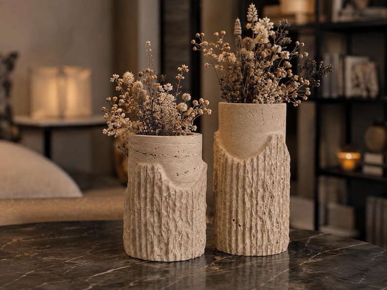 May include: Two cylindrical beige stone vases with a rough, vertically grooved texture on the lower half and a smooth upper section. Each vase contains a bouquet of dried, light-colored flowers with small, delicate, clustered blossoms.