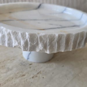 May include: A circular white marble pedestal tray featuring a smooth, polished top surface with grey veining and a rough, chiseled, textured outer edge. The tray is supported by a central cylindrical marble base with matching grey veining.