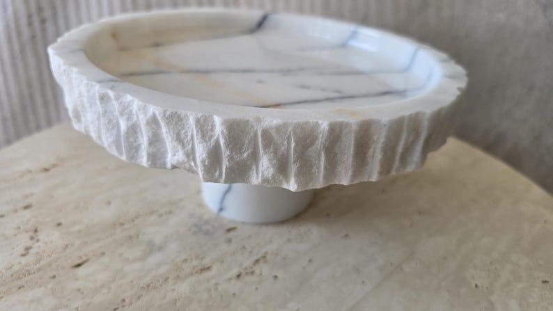 May include: A circular white marble pedestal tray featuring a smooth, polished top surface with grey veining and a rough, chiseled, textured outer edge. The tray is supported by a central cylindrical marble base with matching grey veining.