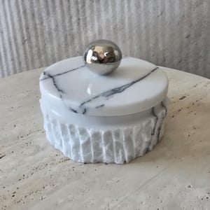 May include: A round white marble container with black veining and a textured, fluted exterior. The lid features a smooth, polished surface and is topped with a reflective, spherical silver-toned metal knob handle centred on the top.