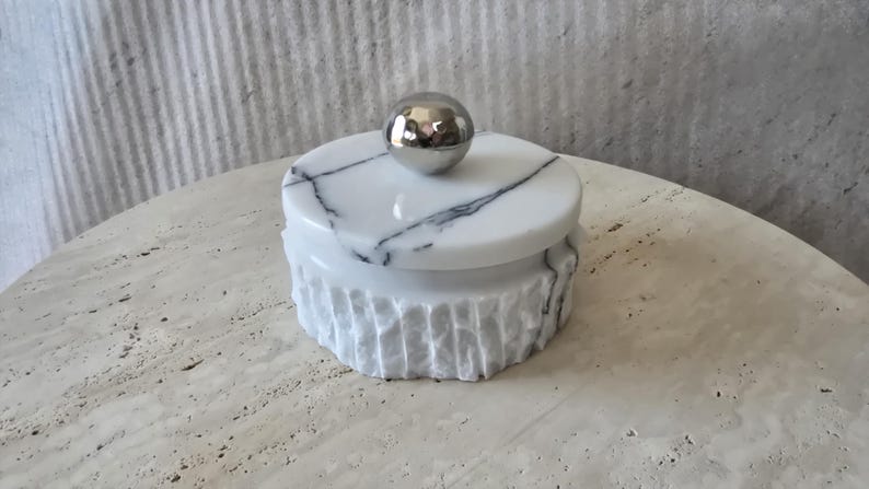 May include: A round white marble container with black veining and a textured, fluted exterior. The lid features a smooth, polished surface and is topped with a reflective, spherical silver-toned metal knob handle centred on the top.