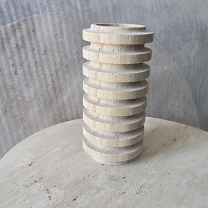 May include: A cylindrical vase crafted from beige travertine stone, featuring a series of stacked, horizontal circular discs that create a ribbed, textured exterior surface with natural porous markings throughout the light tan material.