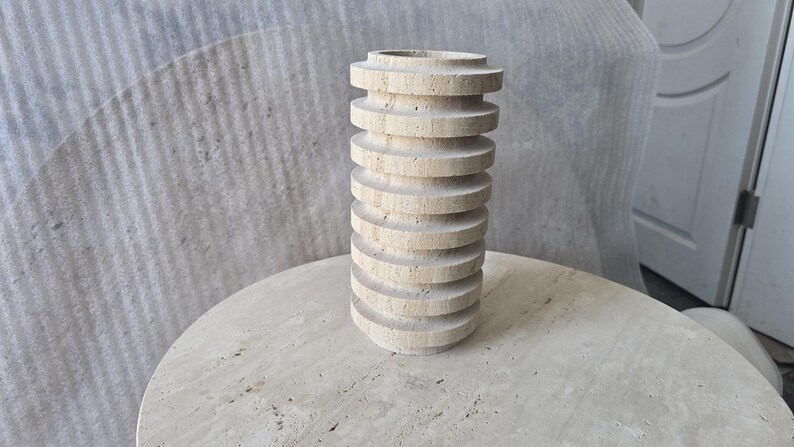 May include: A cylindrical vase crafted from beige travertine stone, featuring a series of stacked, horizontal circular discs that create a ribbed, textured exterior surface with natural porous markings throughout the light tan material.