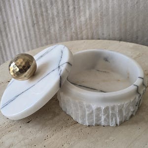 May include: A round white marble container with black veining, featuring a textured, chiseled exterior finish. The lid is smooth and polished, topped with a spherical, hammered gold-tone metal knob handle that serves as a decorative accent.