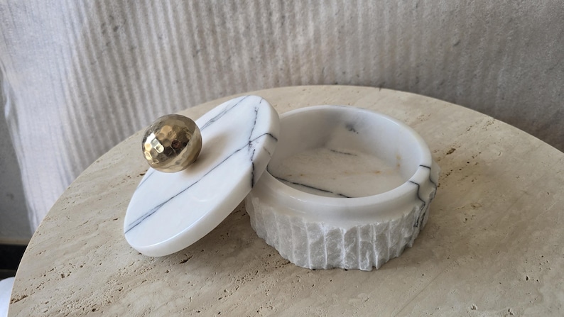 May include: A round white marble container with black veining, featuring a textured, chiseled exterior finish. The lid is smooth and polished, topped with a spherical, hammered gold-tone metal knob handle that serves as a decorative accent.