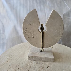 K&ouml;nnte beinhalten: Eine Skulptur aus einem kreisf&ouml;rmigen, beigen Travertinstein mit keilf&ouml;rmigem Ausschnitt, montiert auf einem quadratischen Sockel. Eine kleine, bronzefarbene Metallfigur sitzt in der zentralen &Ouml;ffnung mit nach unten h&auml;ngenden Beinen.