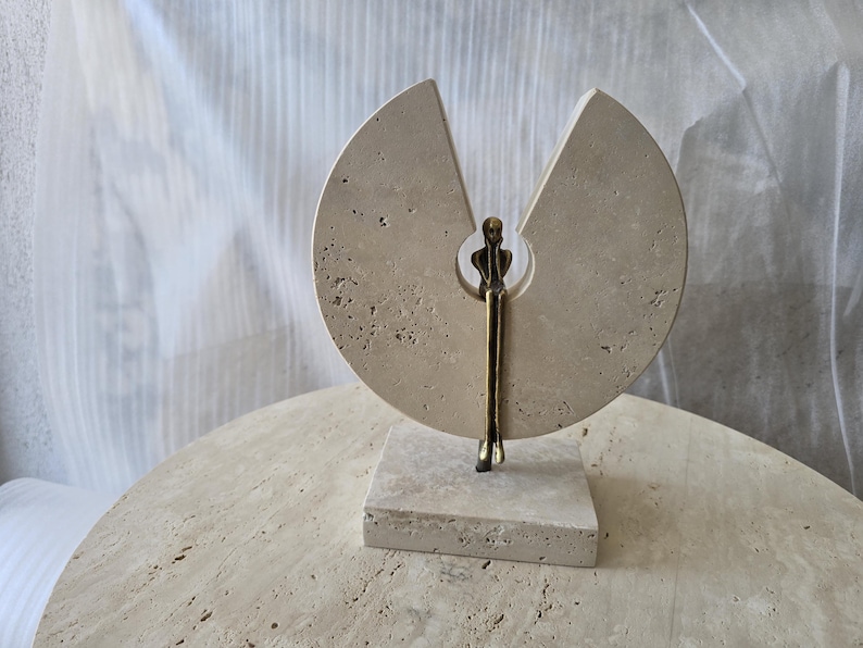K&ouml;nnte beinhalten: Eine Skulptur aus einem kreisf&ouml;rmigen, beigen Travertinstein mit keilf&ouml;rmigem Ausschnitt, montiert auf einem quadratischen Sockel. Eine kleine, bronzefarbene Metallfigur sitzt in der zentralen &Ouml;ffnung mit nach unten h&auml;ngenden Beinen.