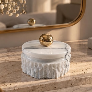 May include: A round white marble storage box with a textured, chiseled exterior and a smooth lid. The lid features a polished, reflective gold-tone spherical knob handle. The marble displays natural grey veining throughout the stone surface.