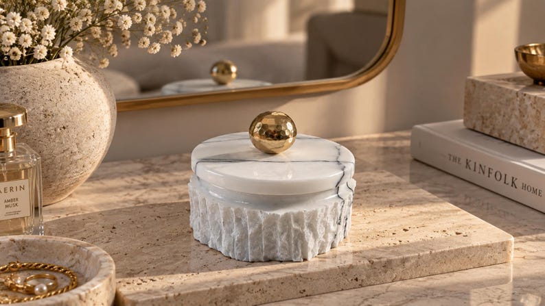 May include: A round white marble storage box with a textured, chiseled exterior and a smooth lid. The lid features a polished, reflective gold-tone spherical knob handle. The marble displays natural grey veining throughout the stone surface.
