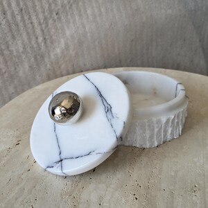 May include: A round white marble container with black veining and a textured exterior. The lid features a smooth, polished surface with a central, spherical, reflective silver-toned metal knob, providing a contrast to the stone's natural finish.