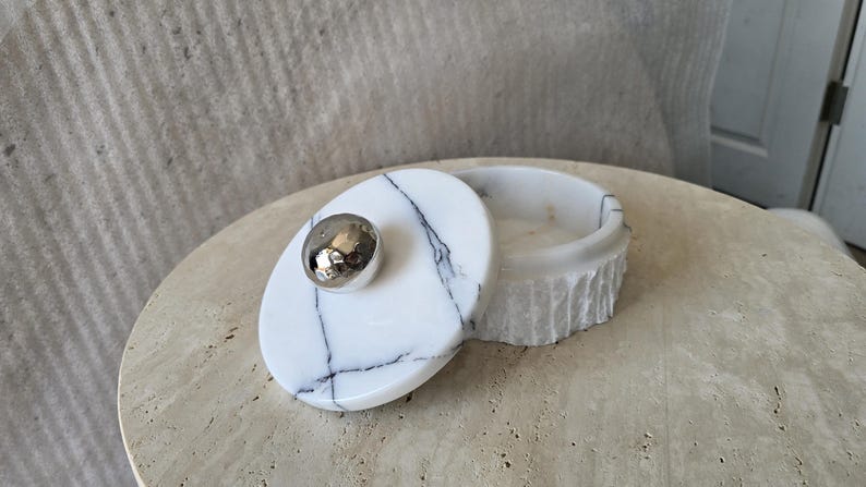 May include: A round white marble container with black veining and a textured exterior. The lid features a smooth, polished surface with a central, spherical, reflective silver-toned metal knob, providing a contrast to the stone's natural finish.