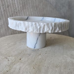 May include: A white marble pedestal stand featuring a smooth, polished top surface with subtle grey veining and a textured, chiseled outer rim. The base is a smooth, cylindrical column of matching white marble with grey veining throughout the stone.
