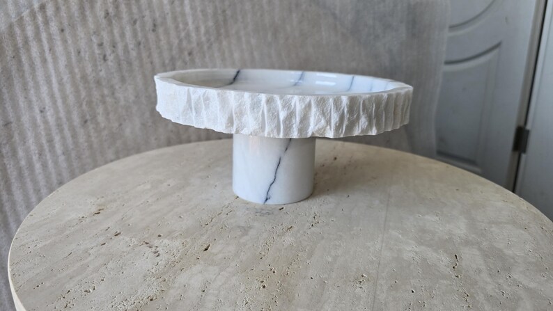May include: A white marble pedestal stand featuring a smooth, polished top surface with subtle grey veining and a textured, chiseled outer rim. The base is a smooth, cylindrical column of matching white marble with grey veining throughout the stone.
