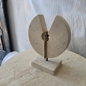 K&ouml;nnte beinhalten: Eine Skulptur bestehend aus einer kreisf&ouml;rmigen Scheibe aus beigem, por&ouml;sem Stein mit einer keilf&ouml;rmigen Aussparung. Eine kleine, metallisch bronzefarbene Figur sitzt in der zentralen &Ouml;ffnung mit herabh&auml;ngenden Beinen auf einem Steinsockel.