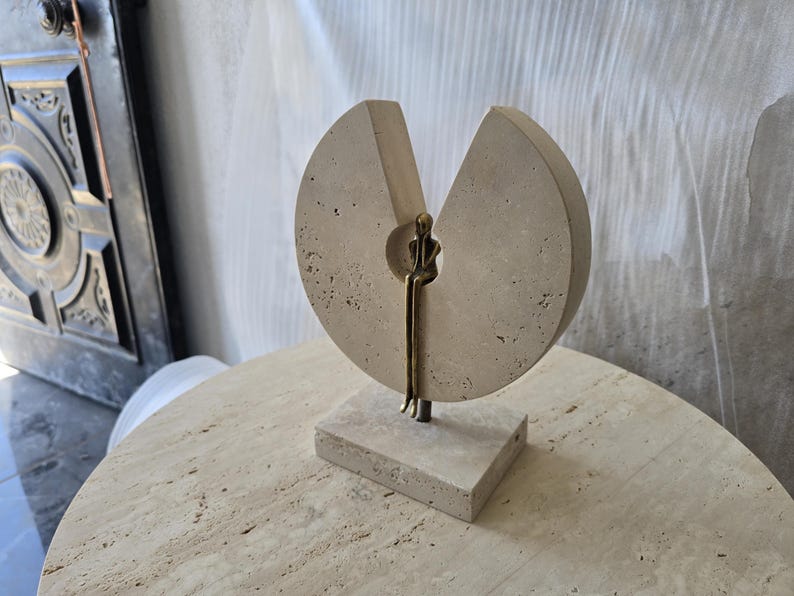 K&ouml;nnte beinhalten: Eine Skulptur bestehend aus einer kreisf&ouml;rmigen Scheibe aus beigem, por&ouml;sem Stein mit einer keilf&ouml;rmigen Aussparung. Eine kleine, metallisch bronzefarbene Figur sitzt in der zentralen &Ouml;ffnung mit herabh&auml;ngenden Beinen auf einem Steinsockel.