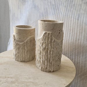 May include: Two cylindrical beige travertine stone vases with a porous, natural texture. Each vase features a smooth upper rim and a lower section with vertical, chiseled grooves that create a rugged, uneven surface appearance throughout.