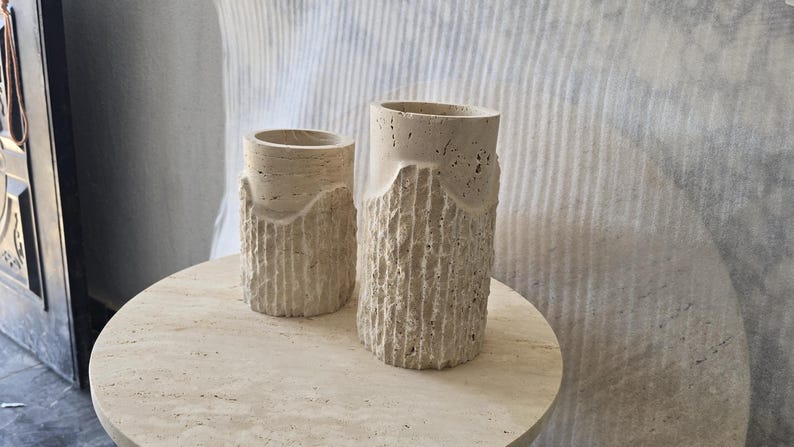 May include: Two cylindrical beige travertine stone vases with a porous, natural texture. Each vase features a smooth upper rim and a lower section with vertical, chiseled grooves that create a rugged, uneven surface appearance throughout.